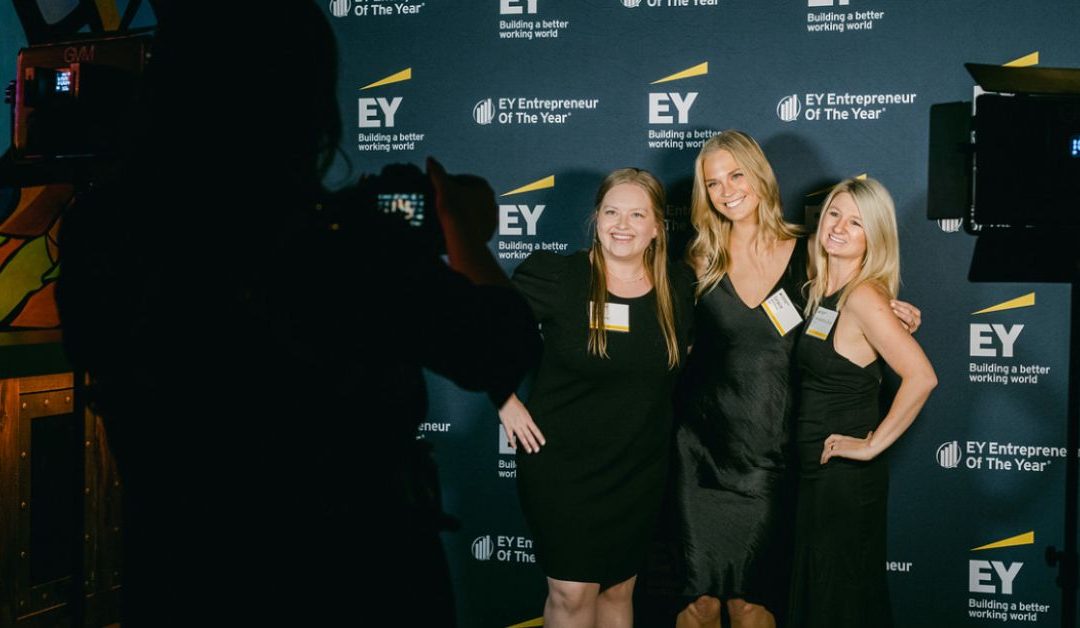 Three ladies stand and get their picture taken in front of a branded backdrop