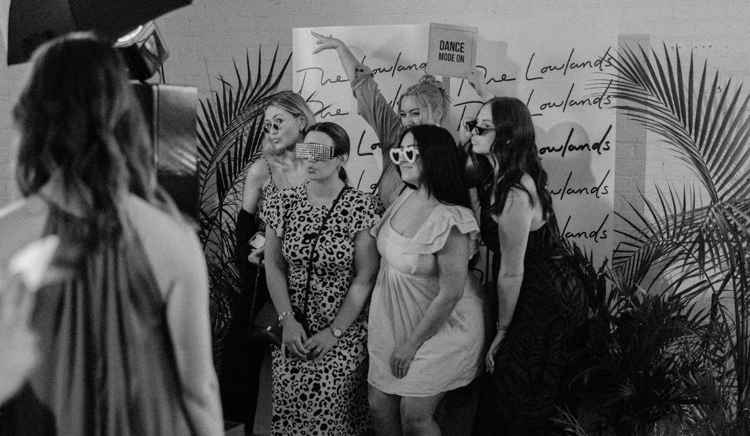 Five women posing together in front of an event backdrop framed by decorative palm leaves. Several wear novelty sunglasses and strike playful poses.