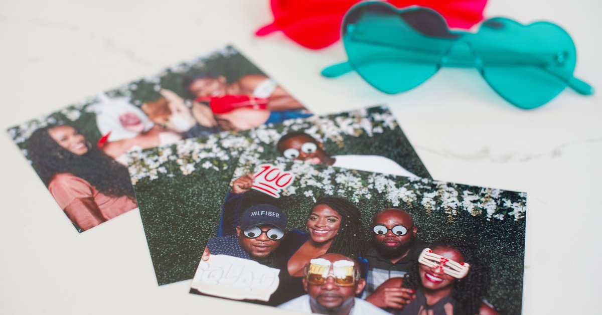 Three printed photo booth pictures on a white surface alongside heart-shaped sunglasses. The photos show groups of people smiling and wearing photo booth accessories.