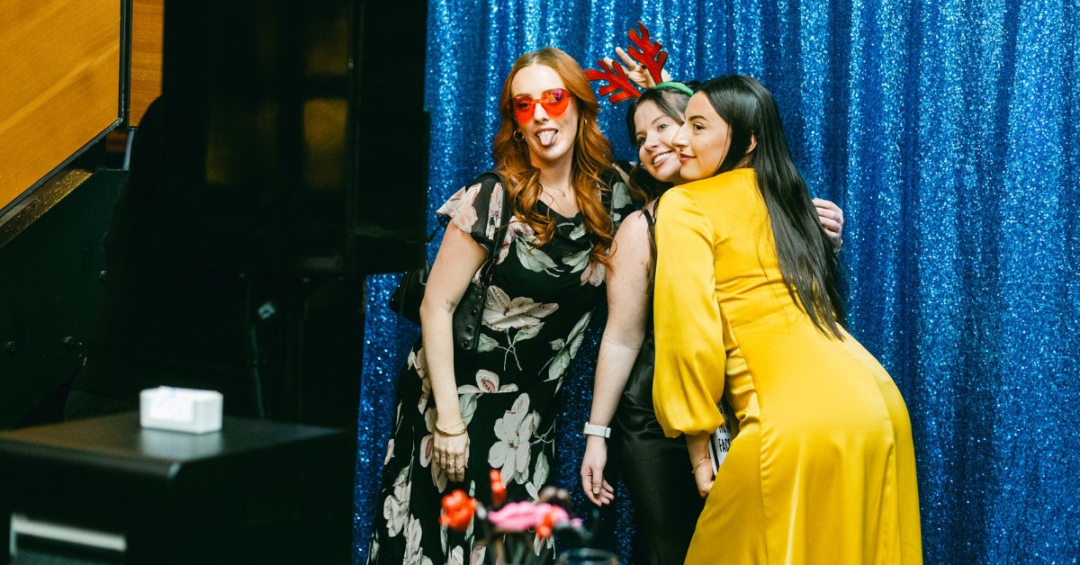 Three women posing together in a photo booth area with a sparkling blue sequin backdrop. One wears red-tinted glasses, another wears festive antler headband props, and all smile while posing.