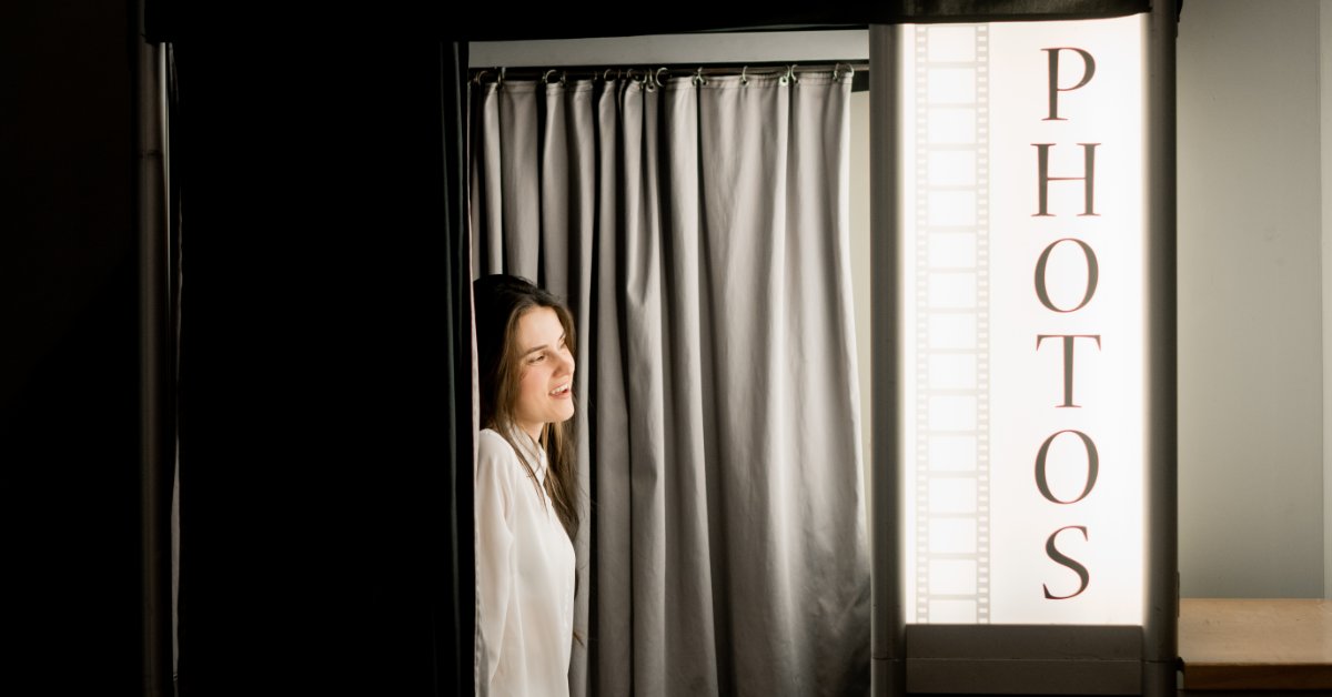 Woman stepping into an enclosed photo booth with black curtains and illuminated “Photos” signage at an event venue.