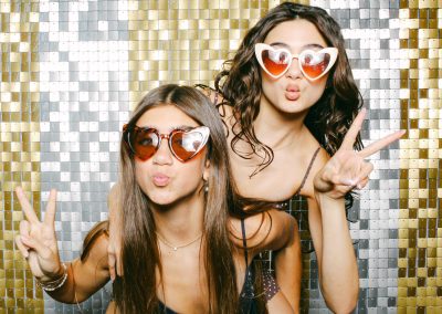 Two teen guests posing with heart sunglasses on a gold sequin backdrop at a mitzvah photo booth