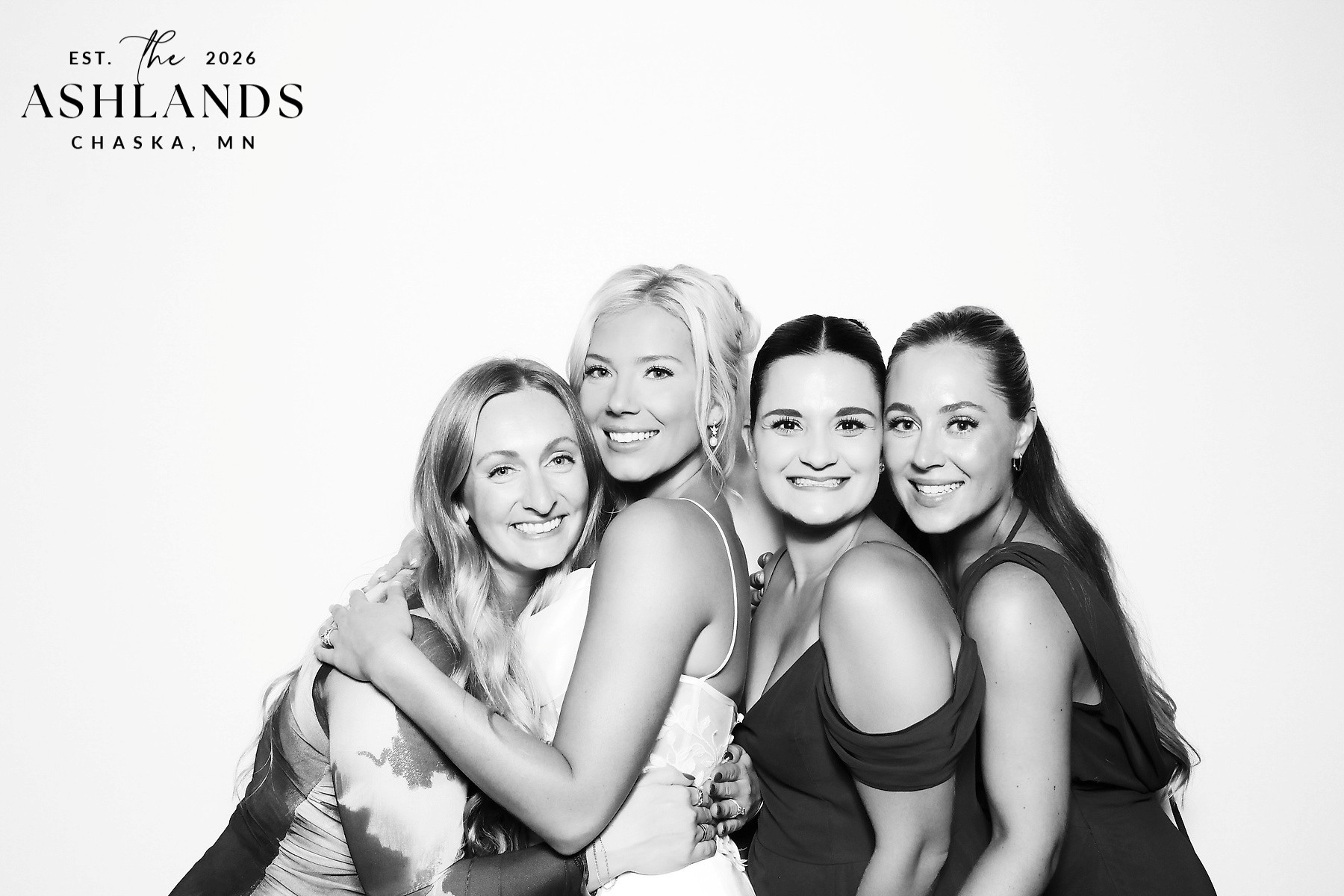 Black and white glam photo booth portrait of four smiling women hugging against a bright white backdrop