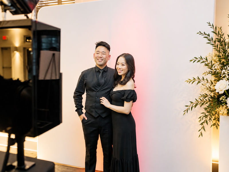 Couple posing at an open style photo booth with a white backdrop and floral arrangement