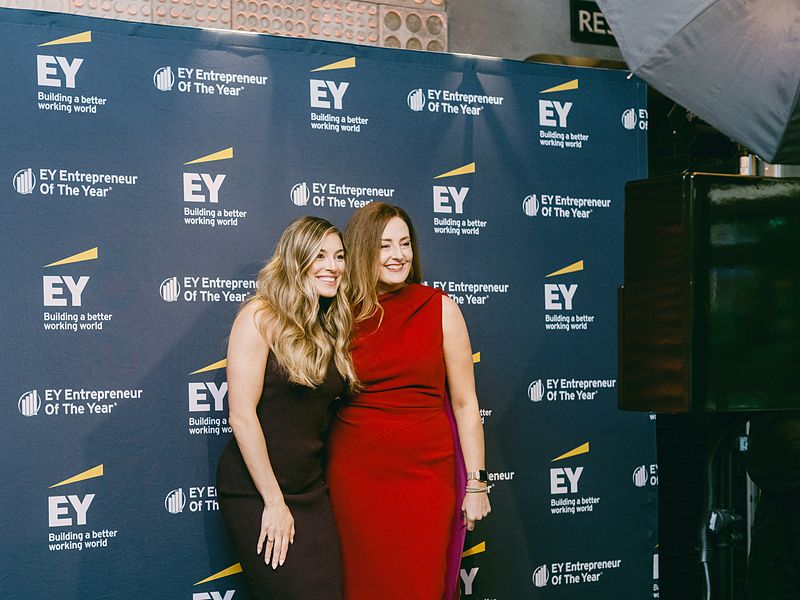Two guests in evening wear at a branded step and repeat backdrop at an awards gala