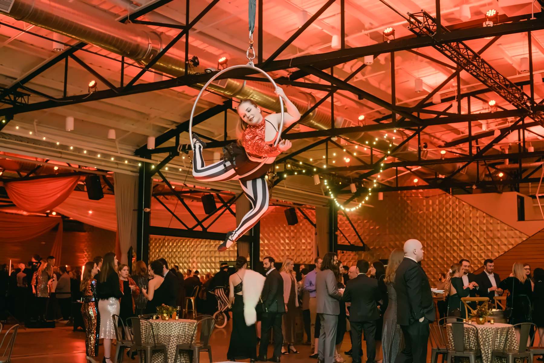 Aerialist on a hoop above a packed corporate party in a warehouse venue lit red and pink with string lights