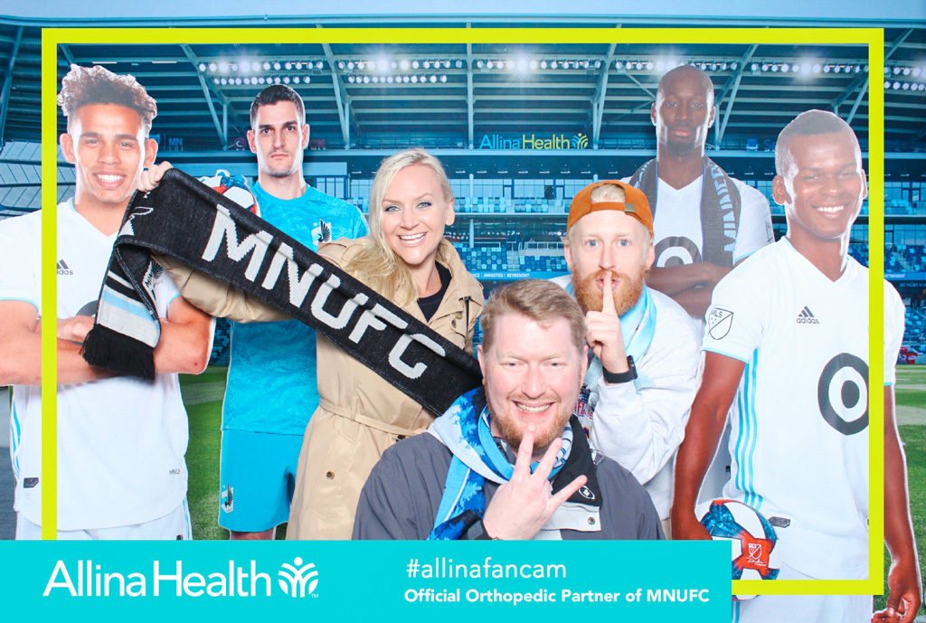 Minnesota United fan cam print with fans posing among life size player cutouts, Allina Health branding