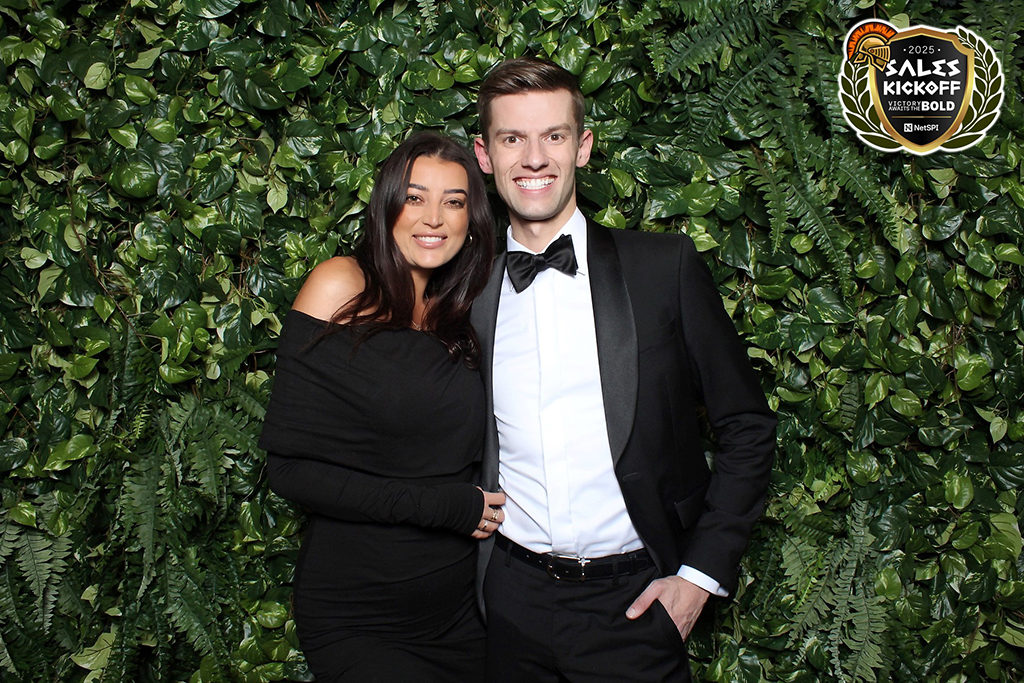 Couple in black tie posing against a green hedge wall at a 2025 sales kickoff